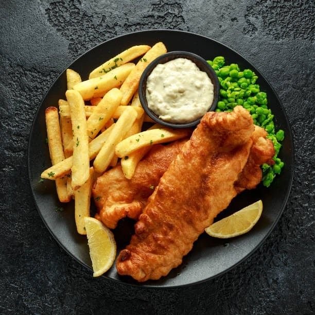 A top-down shot of a serving of fish and chips on a dark plate. Two pieces of golden-brown battered fish are prominently featured alongside a pile of thick-cut french fries. A small black bowl of white tartar sauce and a portion of green peas are also on the plate. Two lemon wedges are placed on either side of the fish.